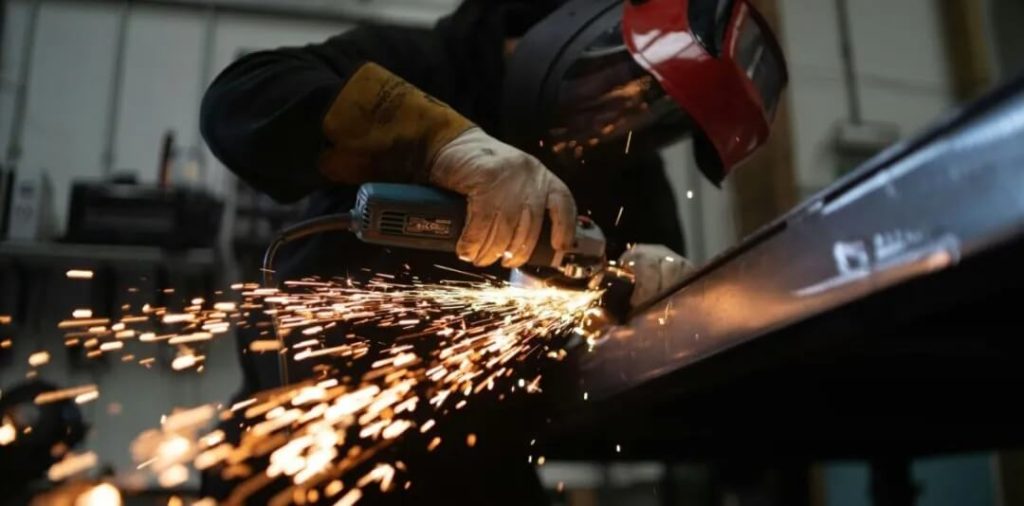 一名工人佩戴防护面罩和手套使用角磨机切割金属,火花四溅,象征工业制造和无损检测中的焊缝打磨场景。