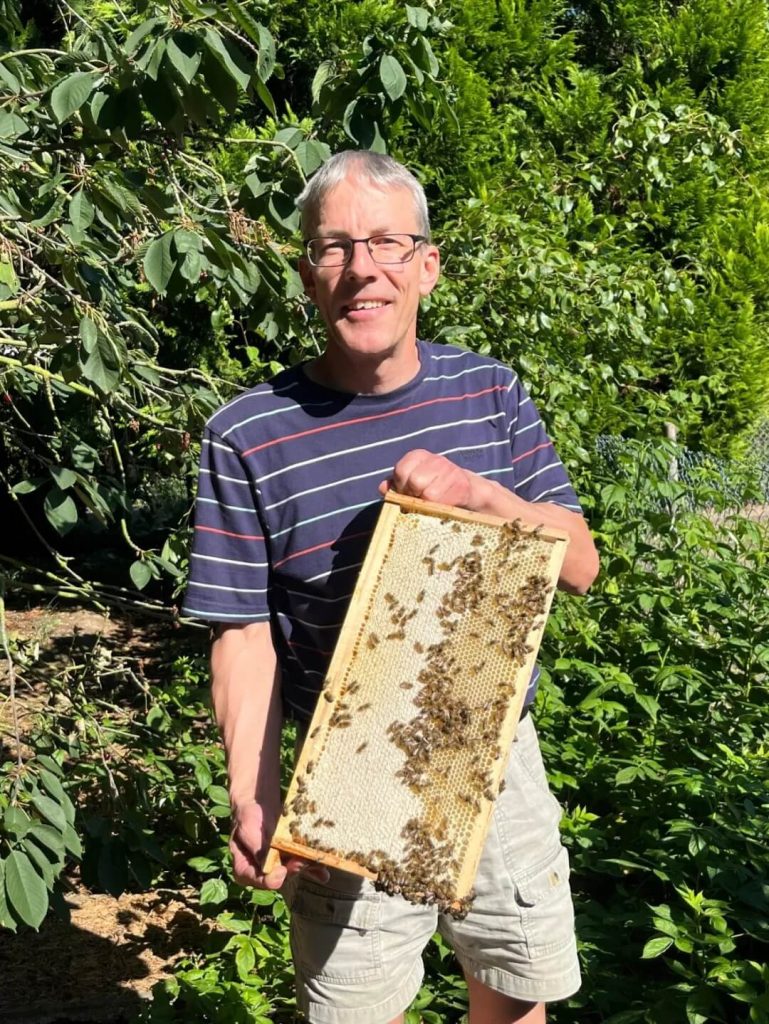 贝克休斯工程师在花园中手持蜂巢检查蜜蜂活动,展示蜜蜂与超声波振动信号之间的自然关联与启发。